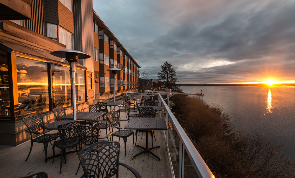 Outdoor patio with tables and chairs, overlooking a river and sunset. The view includes a hotel building, and the sun is setting behind the horizon.